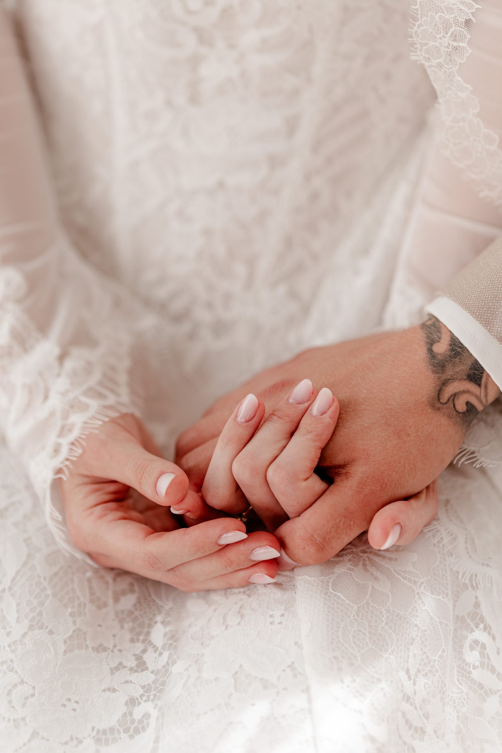 Nahaufnahme während der Trauung: Die Braut im Spitzenkleid hält die tätowierte Hand des Bräutigams in ihrem Schoß