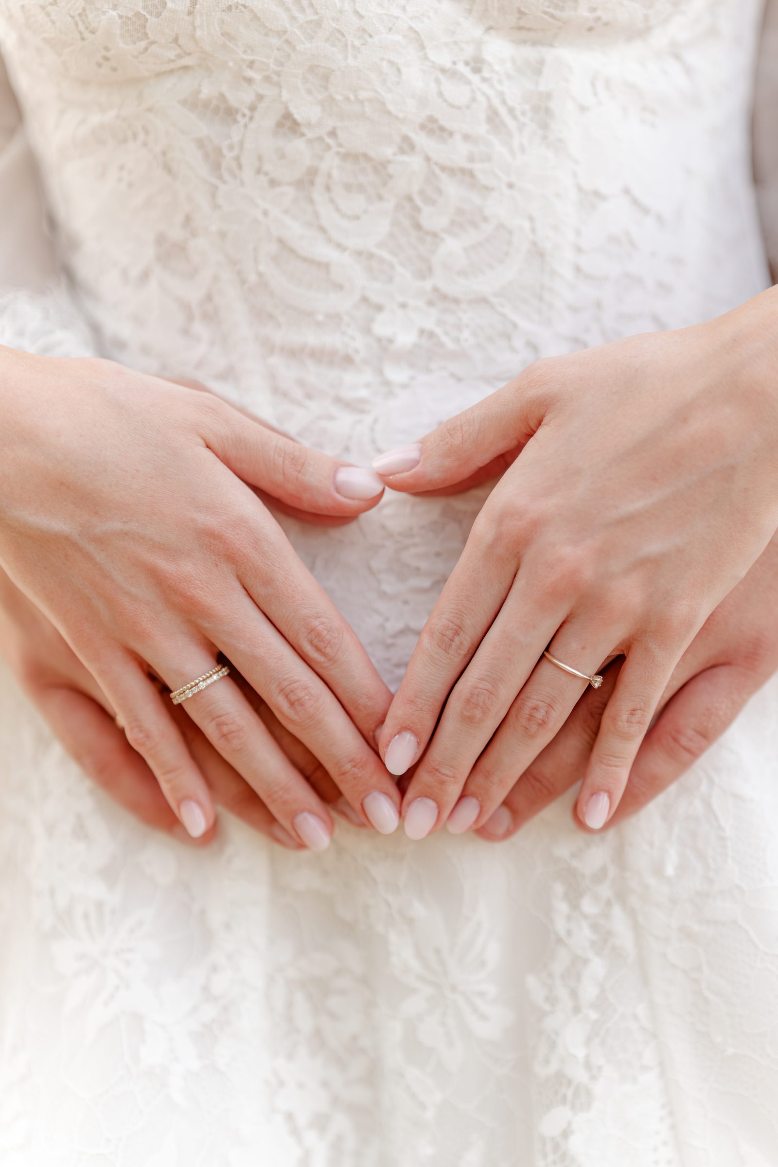 Zwei Hände formen vor dem Spitzenkleid ein Herz, an den Fingern die Eheringe