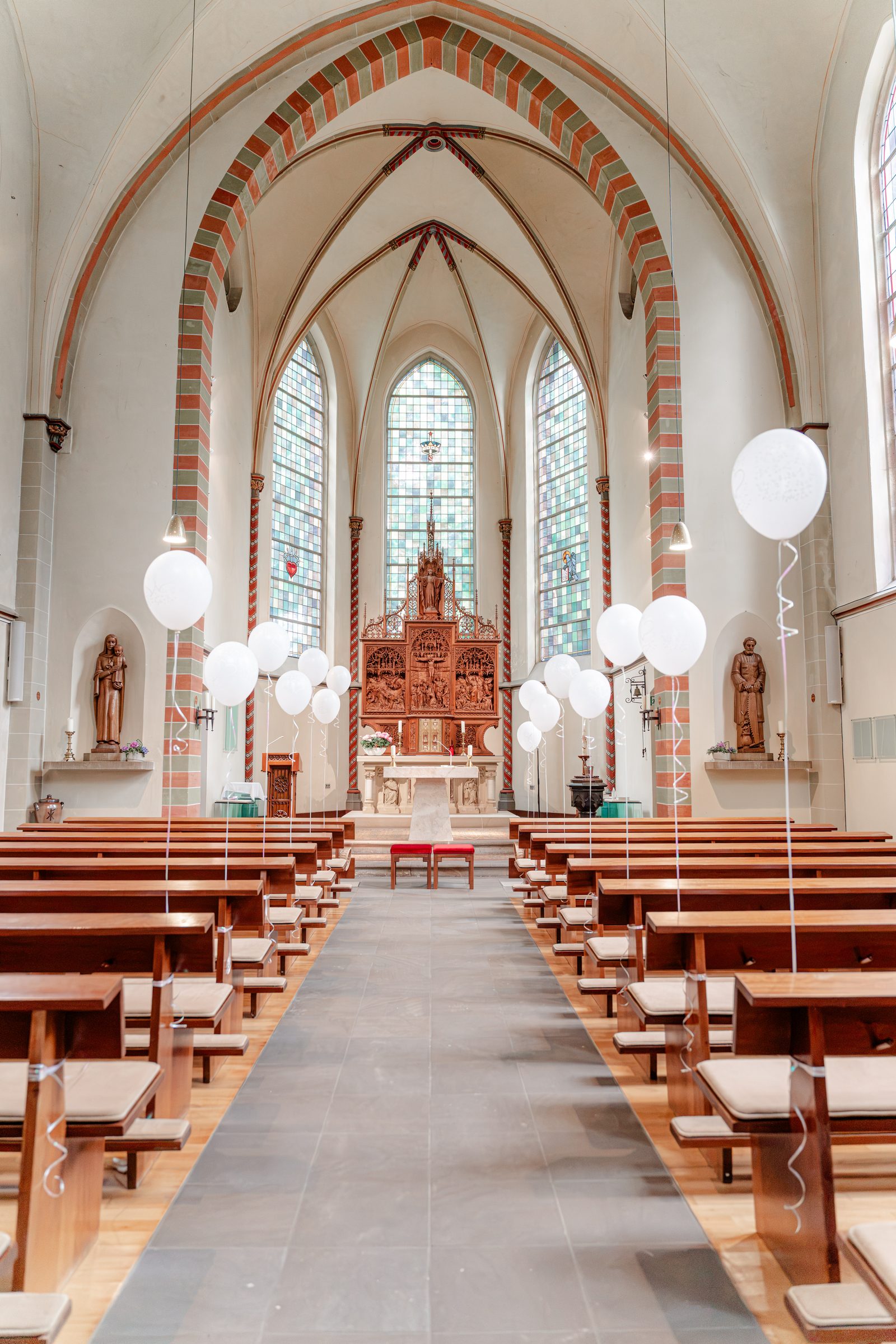 Blick durch den Mittelgang der Herz-Jesu-Kirche zum geschnitzten Altar, an den Bänken weiße Luftballons, Licht fällt durch die bunten Fenster