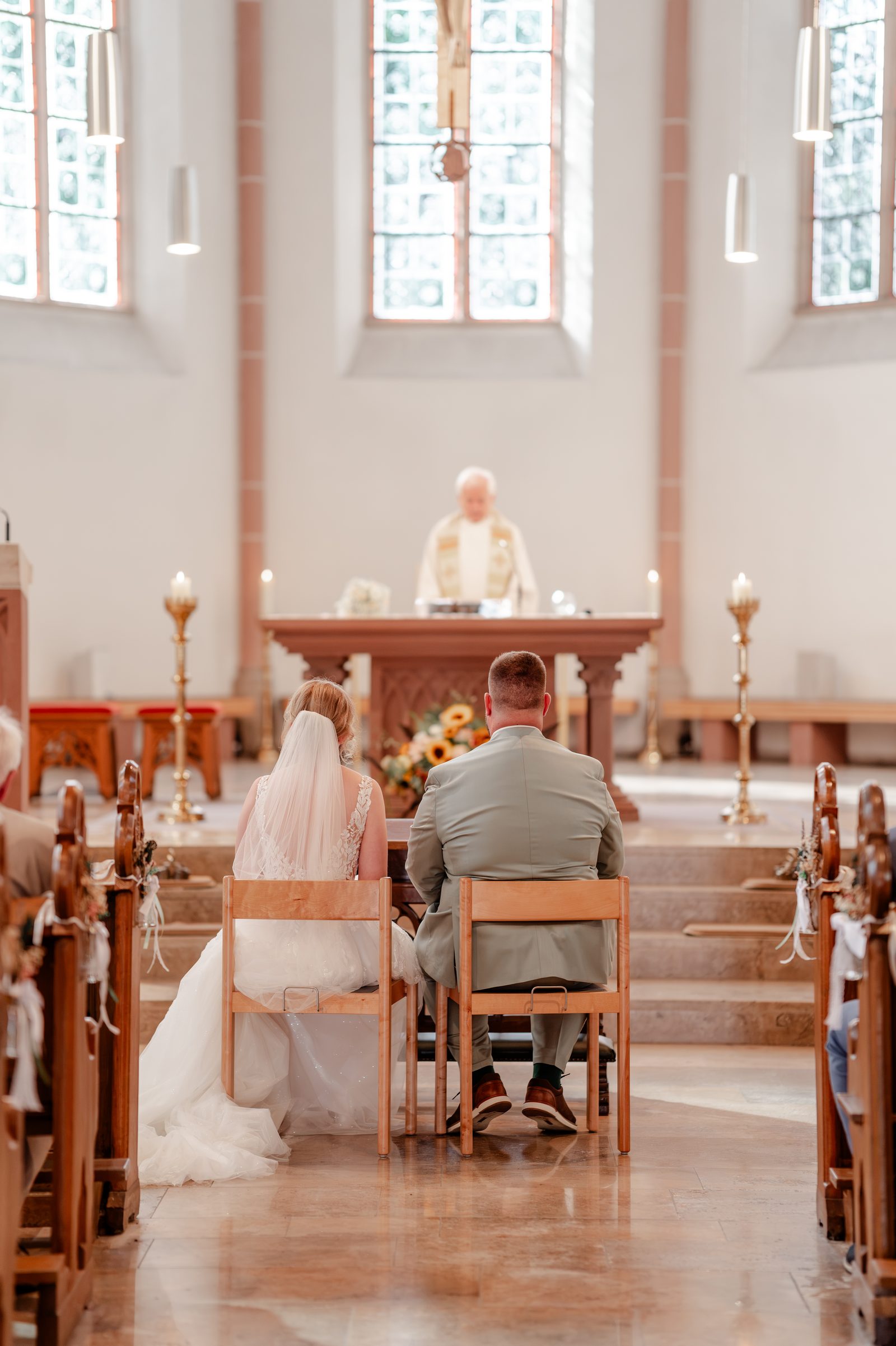 Das Brautpaar sitzt vor dem Altar, dahinter der Pfarrer, Licht fällt durch die hohen Fenster