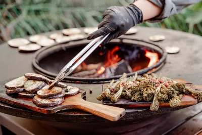 Eine Grillplatte über offenem Feuer, darauf Gemüse, eine Hand mit Grillzange wendet es