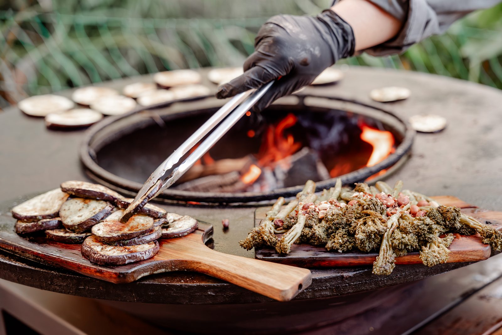 Eine Grillplatte über offenem Feuer, darauf Gemüse, eine Hand mit Grillzange wendet es