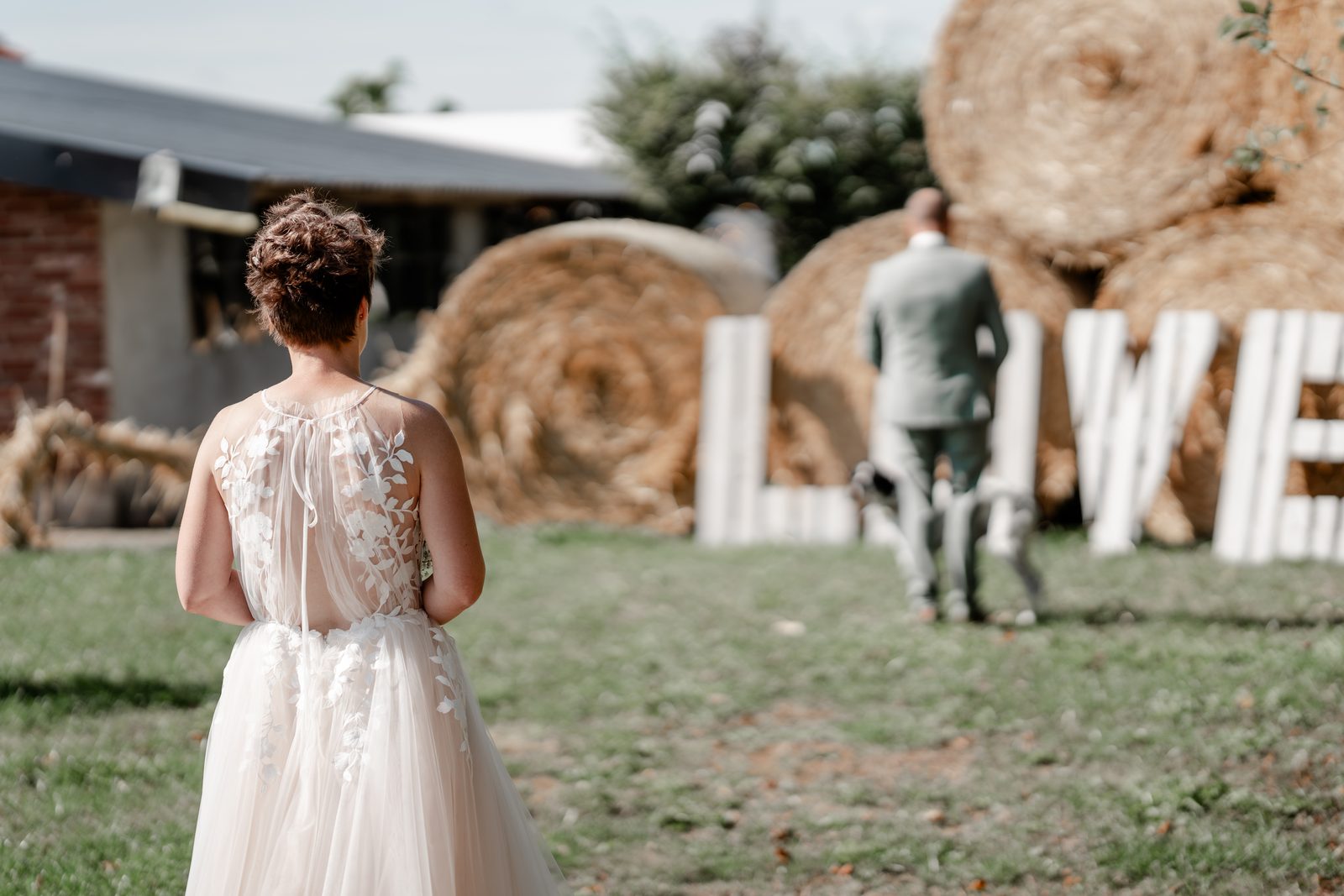 First Look auf dem Hof: Die Braut geht über die Wiese auf den Bräutigam zu, der mit dem Rücken zu ihr vor Strohballen und großen weißen Holzbuchstaben „LOVE“ wartet