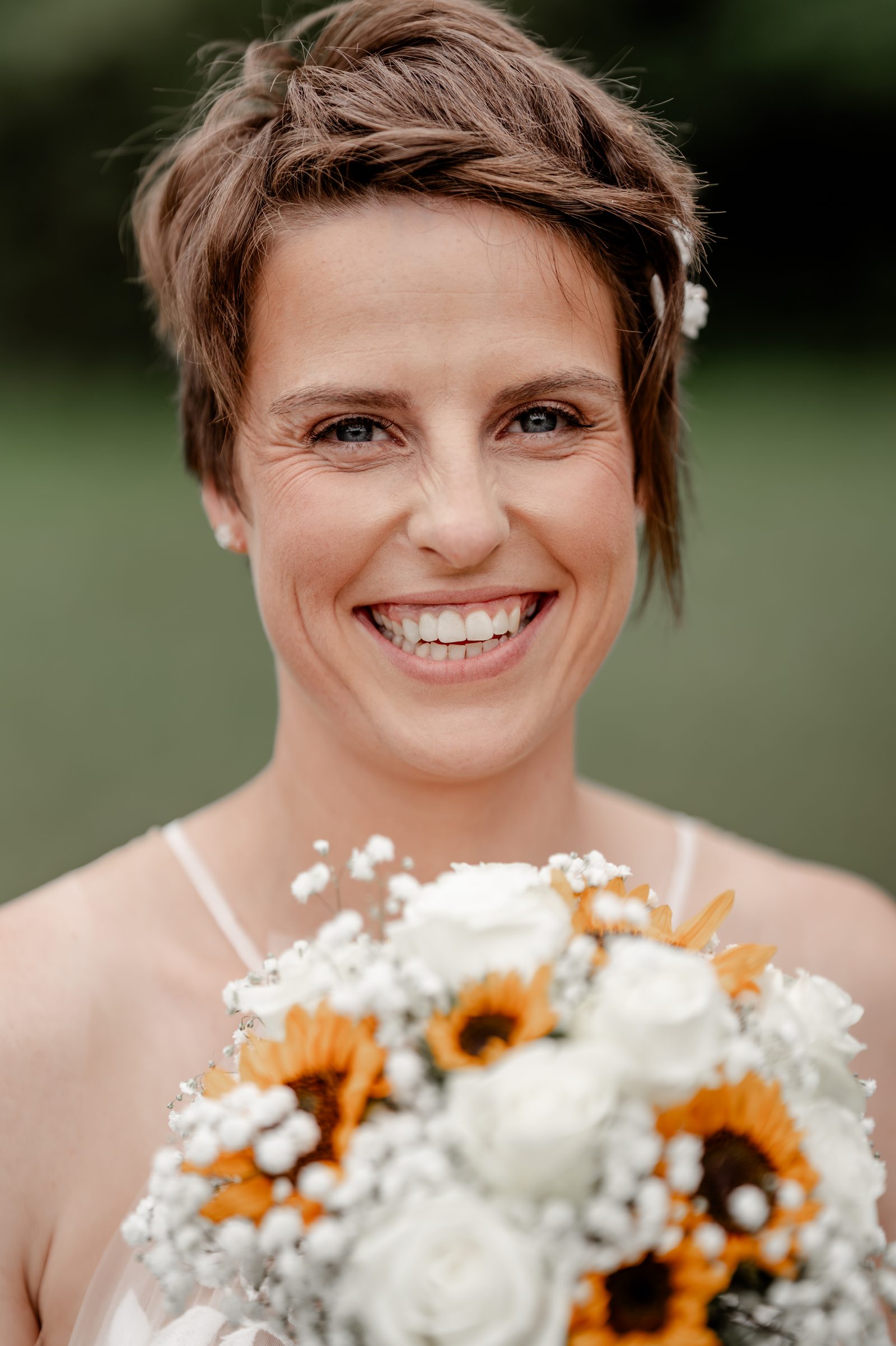 Nahaufnahme der lachenden Braut mit kurzem Haar und Blüte im Haar, vor ihr der Strauß aus Sonnenblumen und weißen Rosen